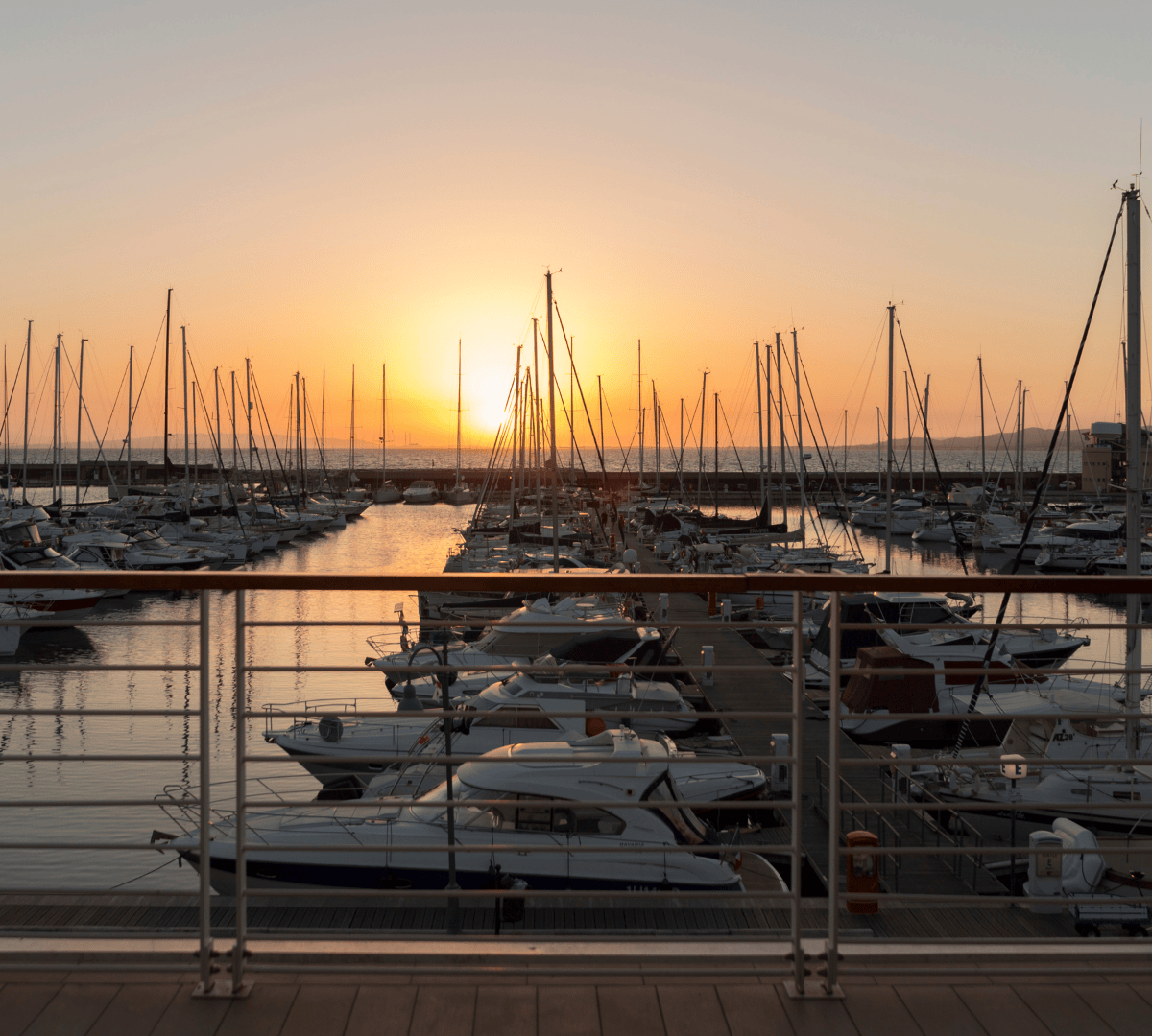 Un tramonto infuocato illumina un porto pieno di barche a vela e yacht, con una ringhiera in primo piano.
