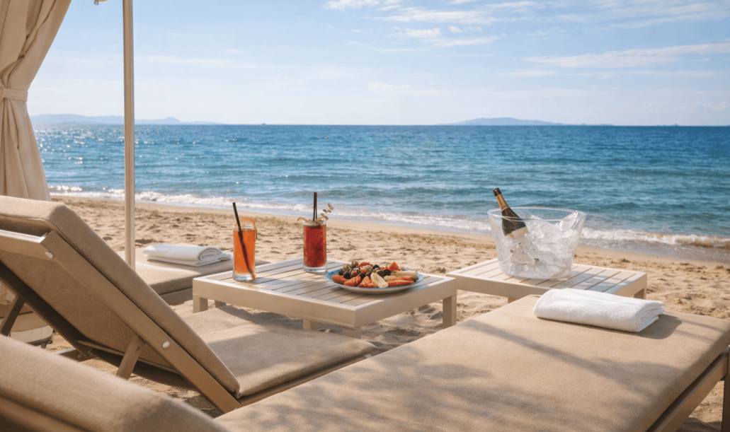 Una veduta in primo piano di eleganti lettini prendisole color sabbia sulla spiaggia della Marina di Scarlino, con drink rinfrescanti, frutta fresca e champagne ghiacciato pronti per un aperitivo in riva al mare sotto un ombrellone chiaro.