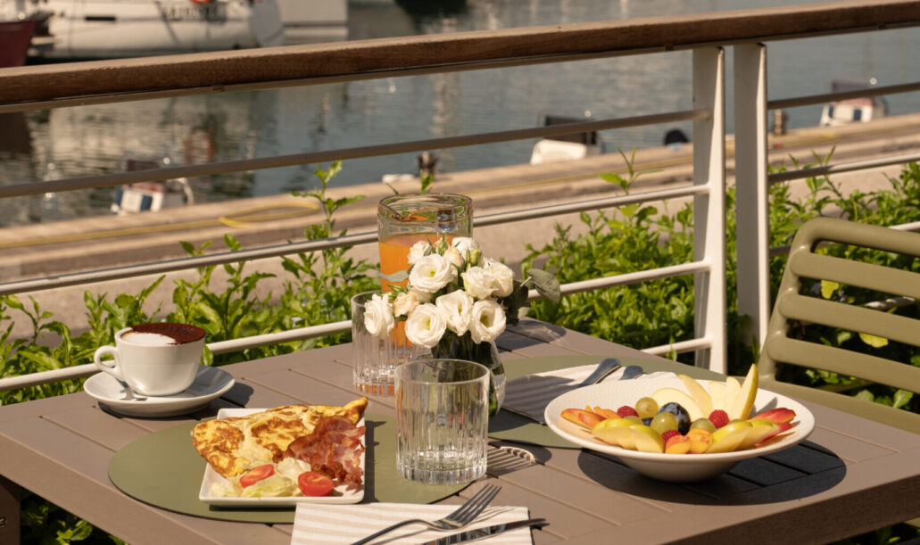Una tavola imbandita per la colazione su una terrazza vista mare al Marina di Scarlino, con caffè, frutta fresca e yacht ormeggiati sullo sfondo.