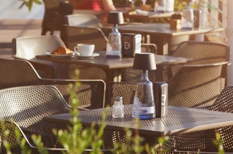 Una terrazza accogliente baciata dalla luce calda del tramonto. In primo piano, tavoli eleganti con lampade di design; sul secondo tavolo si scorgono un caffè e un cornetto pronti per essere gustati, circondati da una vegetazione mediterranea curata.