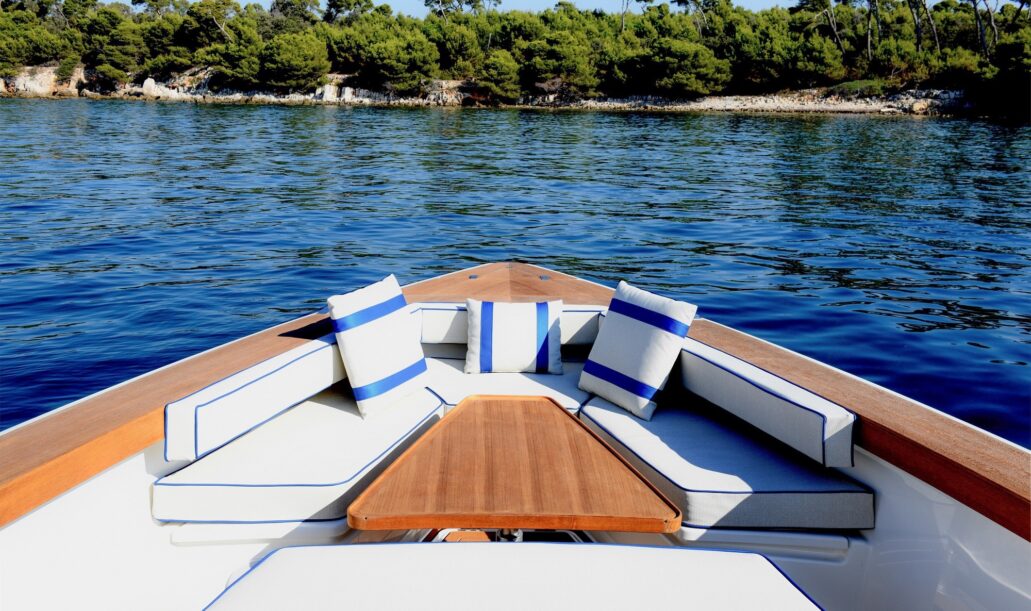 Area relax a prua dello yacht Swan Shadow con cuscineria bianca e blu e tavolino in legno, vista su costa incontaminata della Maremma.