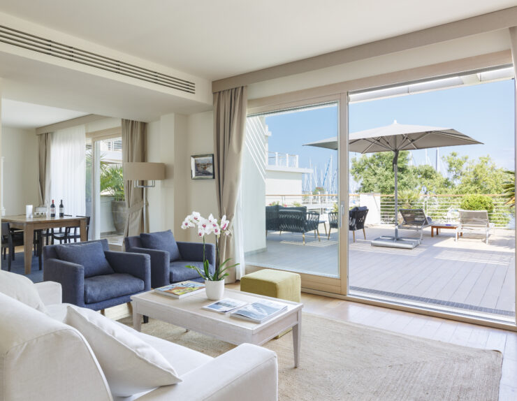 Elegante suite d'hotel con un luminoso soggiorno open space arredato in stile moderno. Si apre su un'ampia terrazza privata pavimentata in legno che offre una vista spettacolare sul porto turistico pieno di barche a vela.