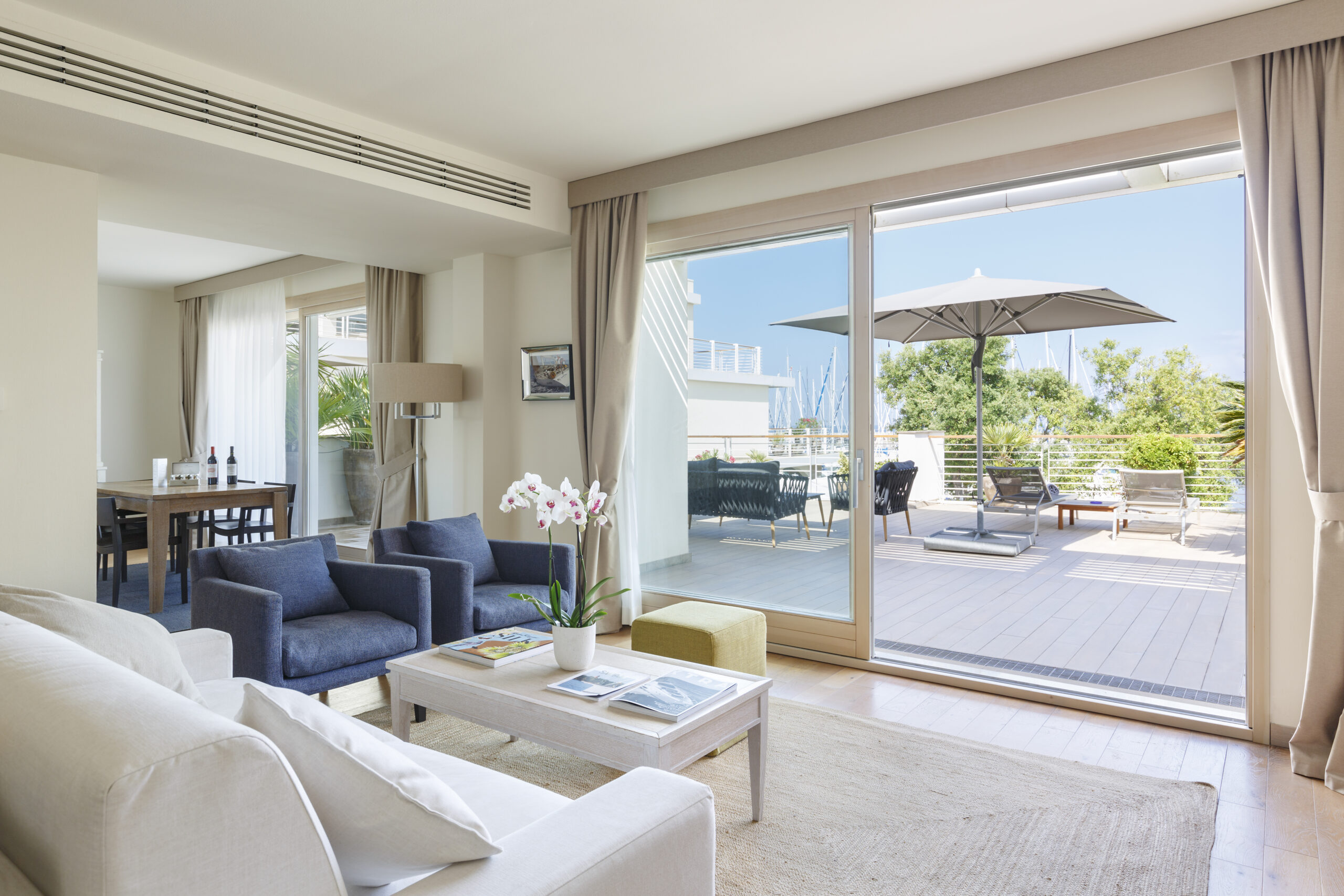 Elegante suite d'hotel con un luminoso soggiorno open space arredato in stile moderno. Si apre su un'ampia terrazza privata pavimentata in legno che offre una vista spettacolare sul porto turistico pieno di barche a vela.