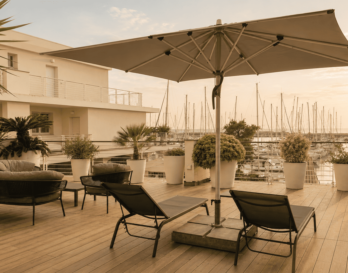 Una terrazza panoramica del Marina di Scarlino Resort con pavimentazione in legno, lettini prendisole, divani scuri e un grande ombrellone centrale, che offre una vista suggestiva sulle barche ormeggiate nella Marina al tramonto.