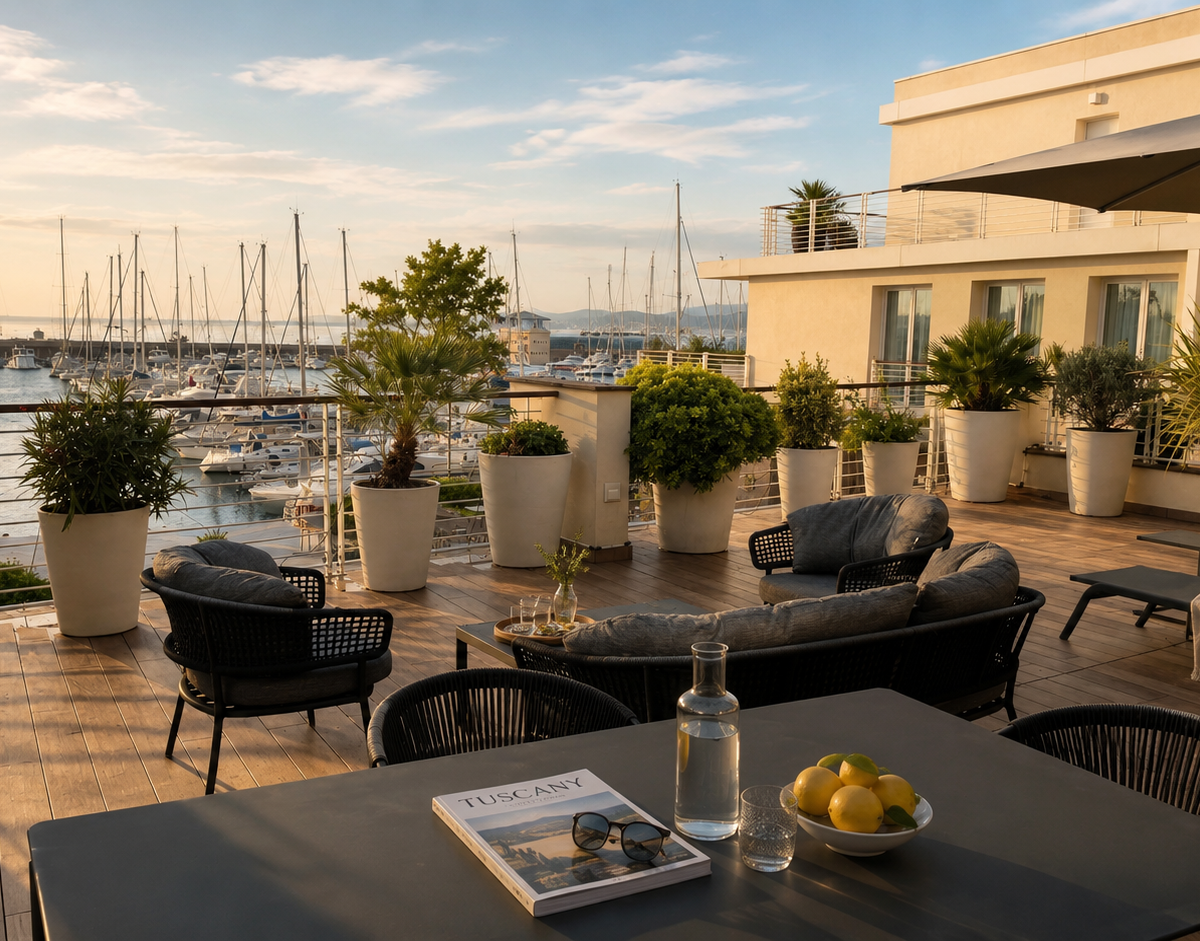 Una spaziosa terrazza arredata del Marina di Scarlino Resort al tramonto, con divani moderni, tavoli e piante in grandi vasi bianchi che si affaccia direttamente sul porto turistico con numerose barche a vela ormeggiate.